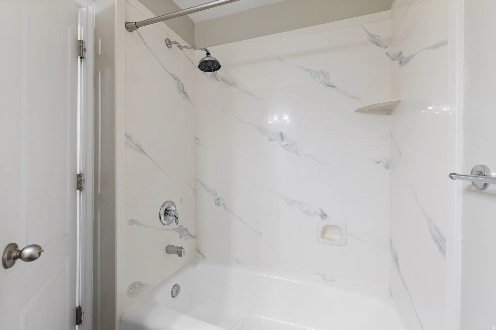 Bathtub and shower combination with a curved shower curtain rod, featuring a white shower surround with grey veining, a built-in corner shelf, and chrome fixtures