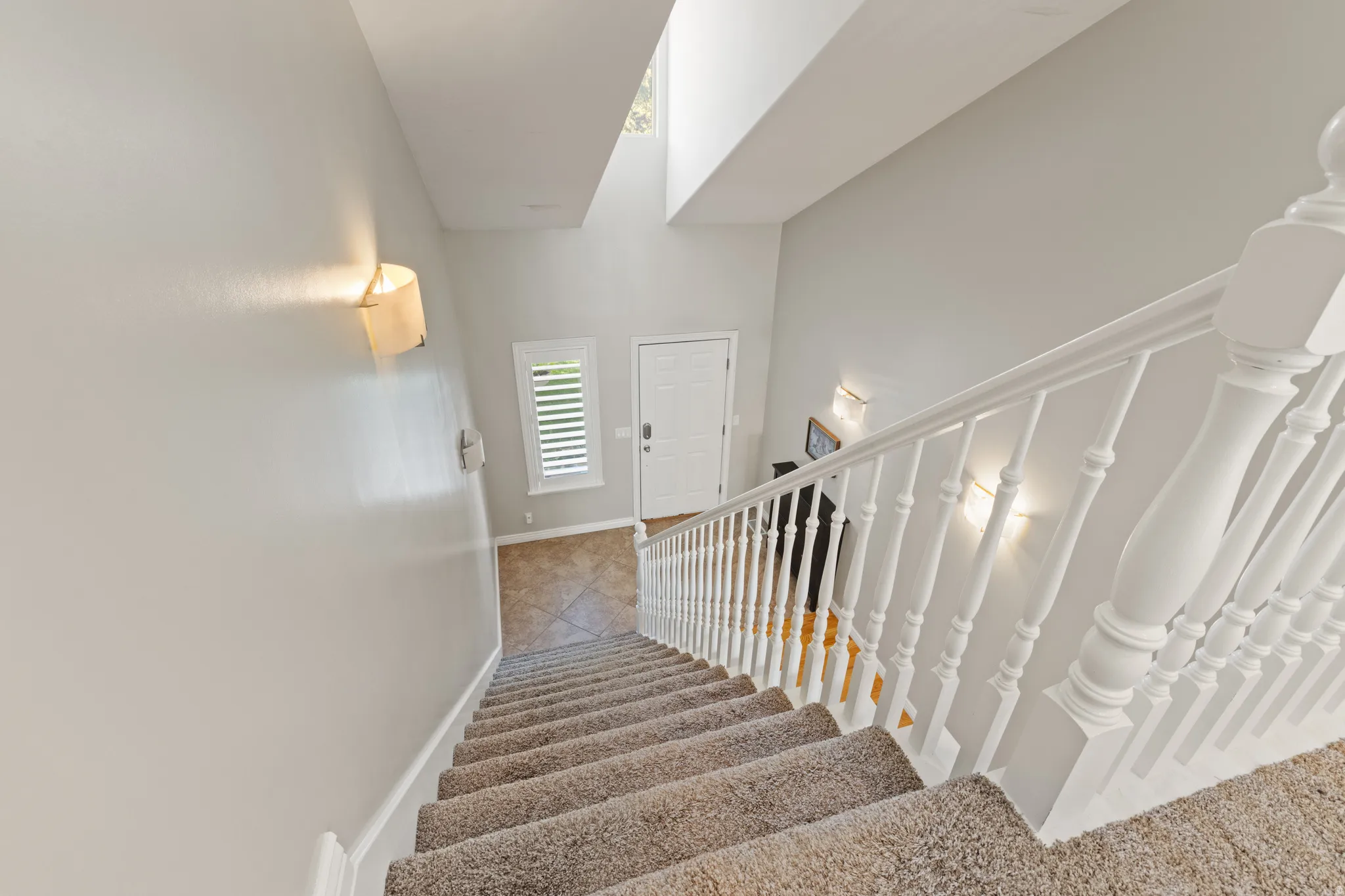 Carpeted staircase with white balusters, leading to a tiled entry with a white door and natural light from a window with white shutters