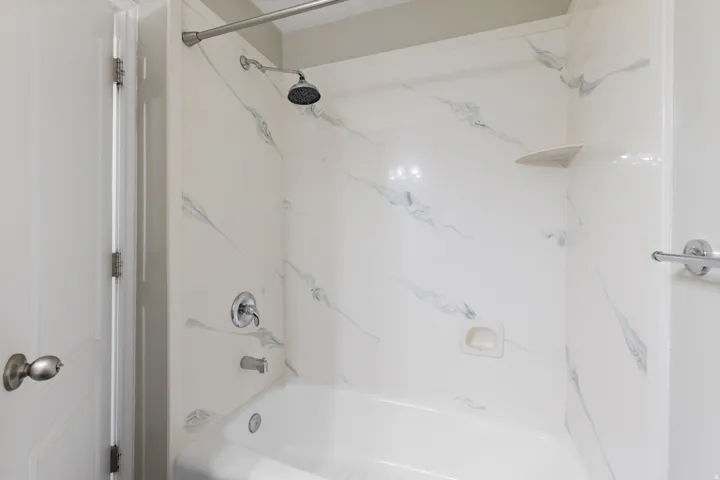 Bathtub and shower combination with a curved shower curtain rod, featuring a white shower surround with grey veining, a built-in corner shelf, and chrome fixtures