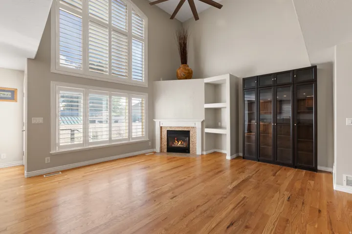 Vaulted ceiling with a ceiling fan, wood-finish flooring, built-in shelving, glass-front cabinetry, and a fireplace with a stone surround
