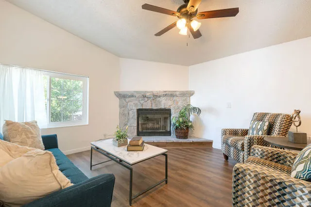 Vaulted ceiling with a ceiling fan, stone-clad corner fireplace with a hearth, wood-finish flooring, and a large window