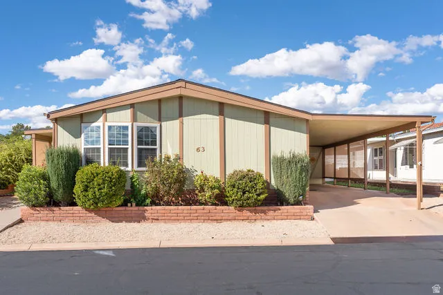 Light green exterior with brown trim, covered carport, brick-lined landscaping, and mature shrubbery