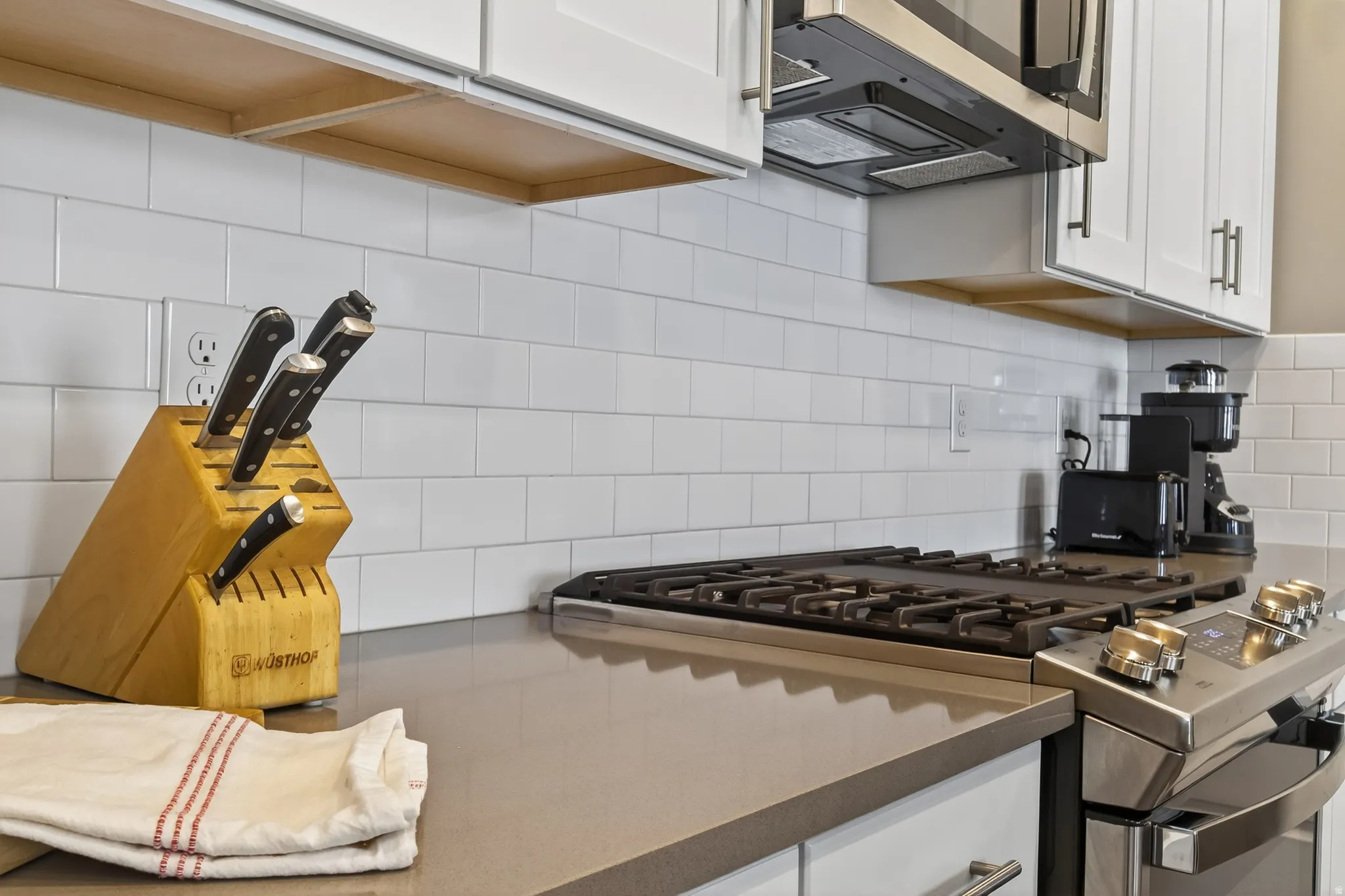 White subway tile backsplash, light-toned countertops, stainless steel range with gas burners, and white upper cabinetry with chrome hardware