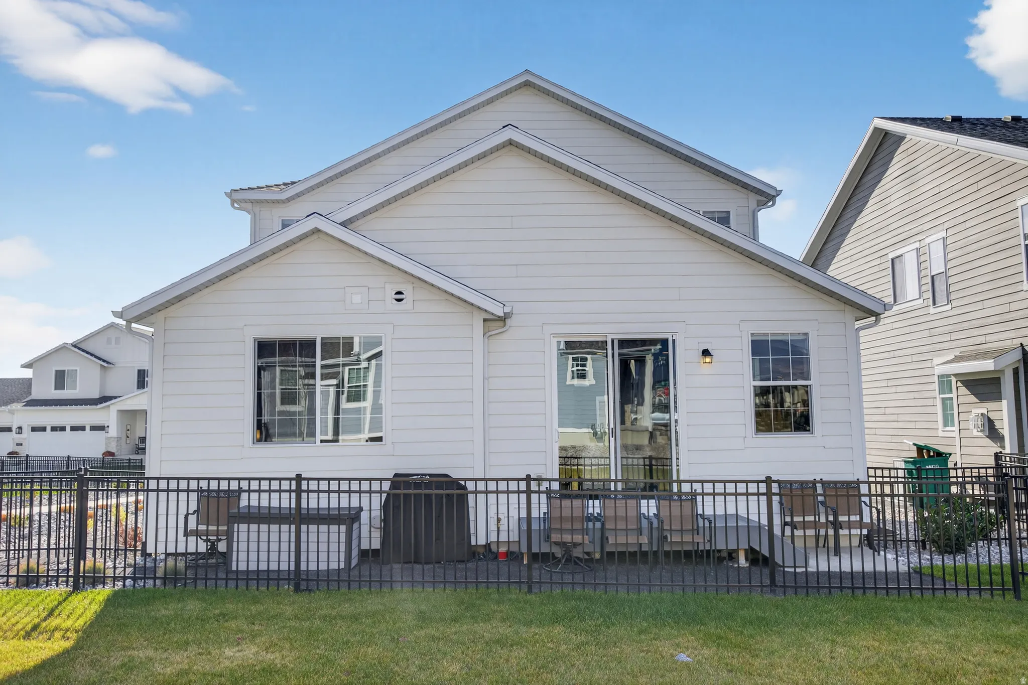 White siding exterior with a pitched roof, featuring a sliding glass patio door, an exterior light fixture, and a black metal fence enclosing a concrete patio