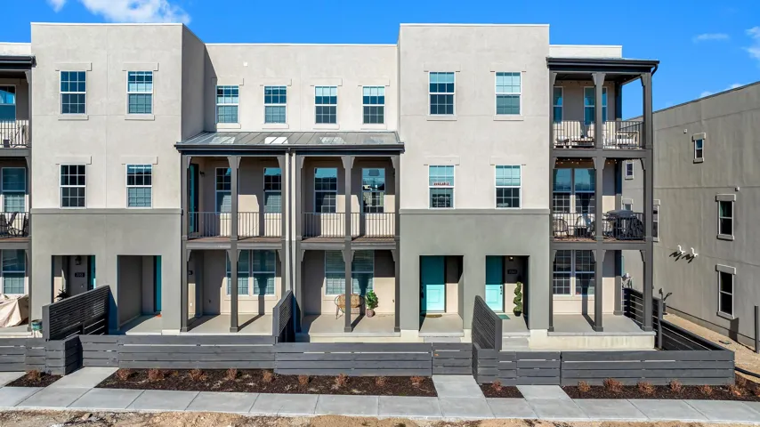 Multi-story townhome with stucco exterior, multiple private balconies with black railings, ground-level patios, and a contemporary fence