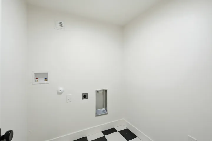 Checkerboard floor tile, white baseboards, wall-mounted electrical outlet, and plumbing hookups
