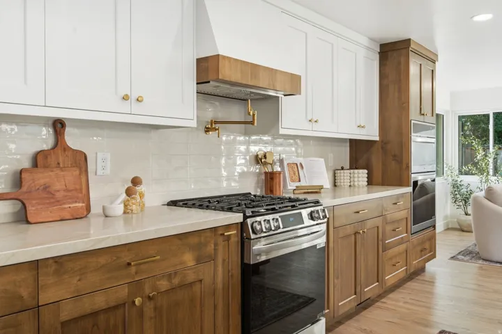 Warm wood-finish flooring with white and wood-finish cabinetry, light stone countertops, a subway tile backsplash, and a stainless steel oven range