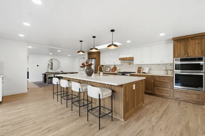 Light wood-finish flooring throughout, kitchen island with seating, stainless steel double ovens, white upper cabinetry, and wood-finish lower cabinetry