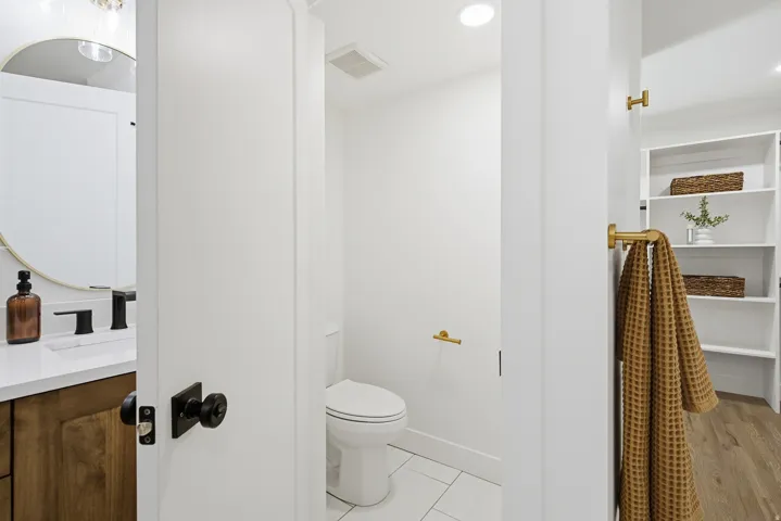 Modern bathroom featuring a white subway tile shower enclosure with frameless glass door, dual-sink vanity with wood-finish cabinetry and brushed gold hardware, white ceramic toilet, light wood-finish flooring, and built-in shelving with woven storage bas