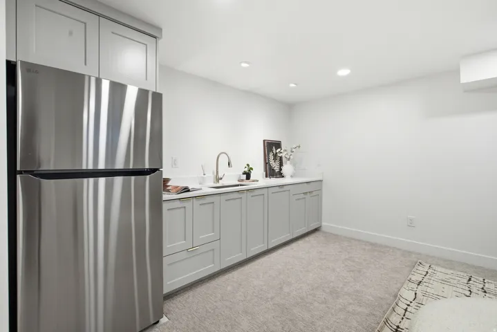 Stainless steel refrigerator, built-in cabinetry with light-colored countertops, undermount sink with gooseneck faucet, light gray shaker-style cabinets, and recessed lighting
