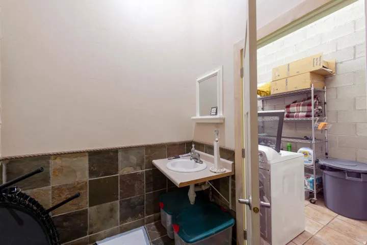 Wall-mounted sink with a countertop, tiled wainscoting, and an adjacent laundry area with a washer and shelving