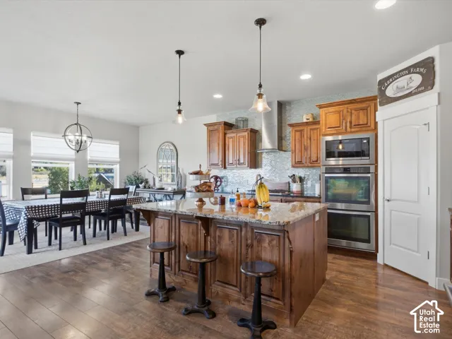 Wood-finish flooring throughout, kitchen island with granite countertop, stainless steel appliances, wood cabinetry, and a decorative tile backsplash