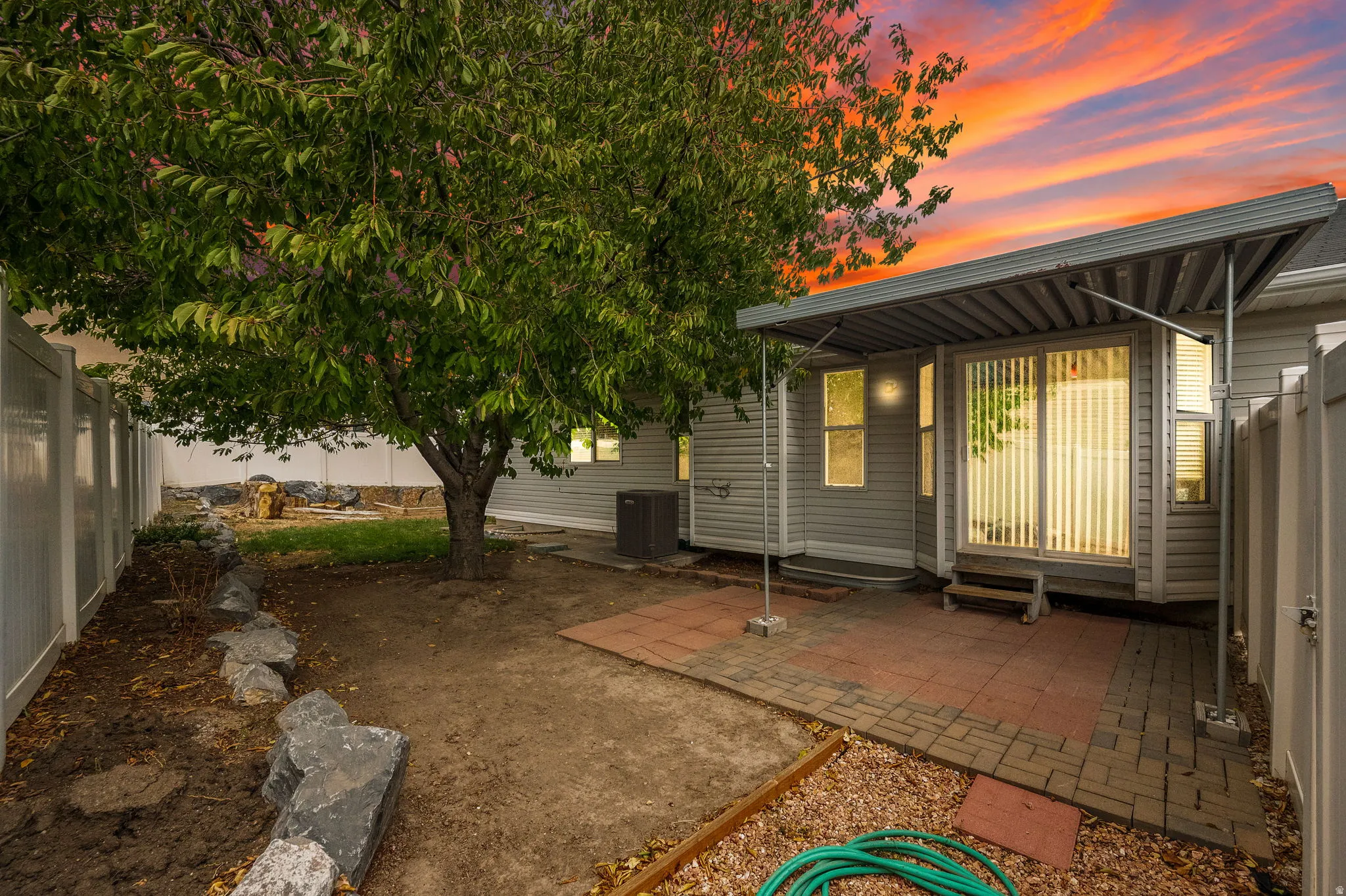Enclosed yard featuring a mature tree, privacy fencing, and a paver patio with a covered awning