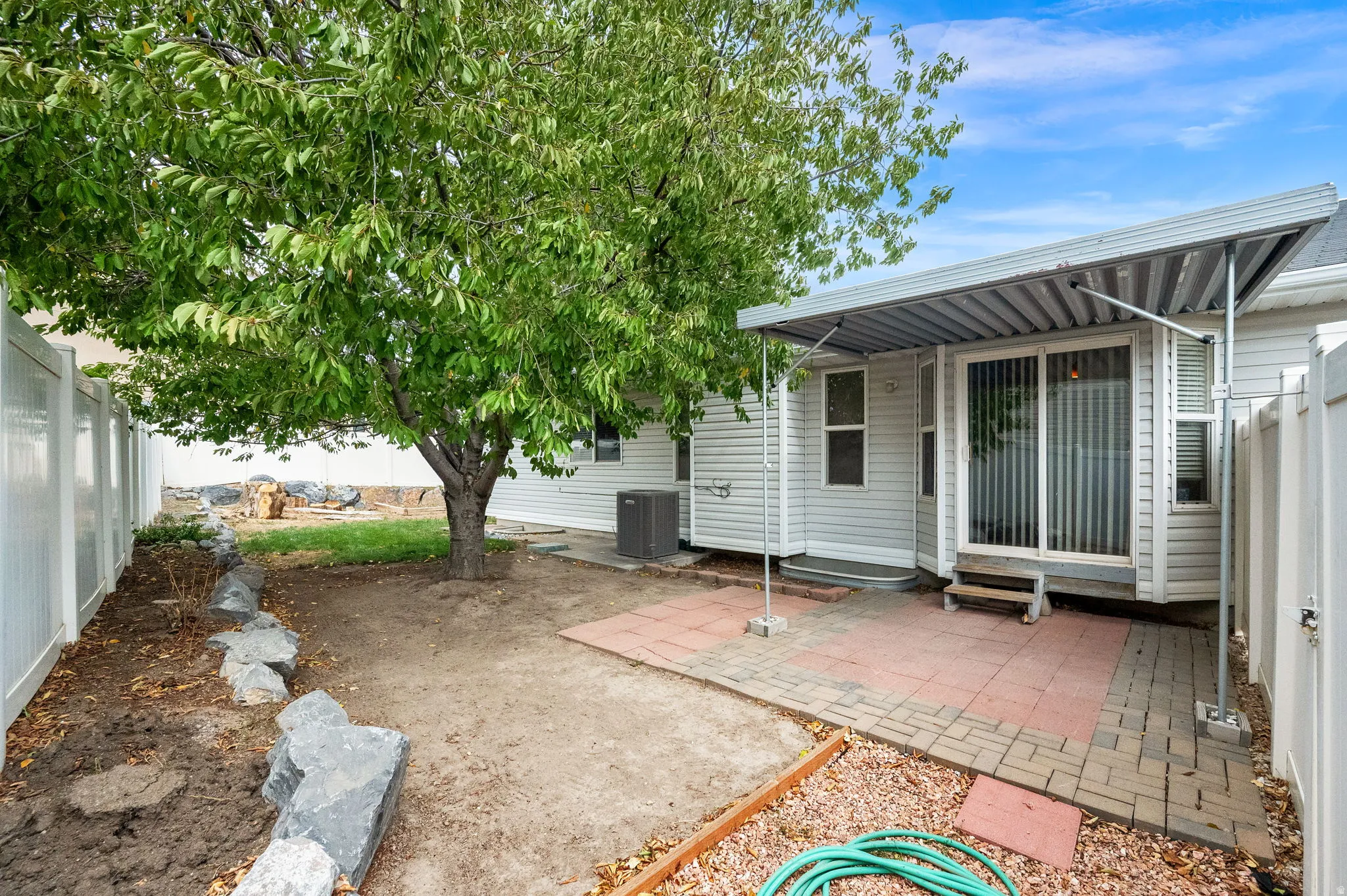 Private backyard with a mature shade tree, white vinyl fencing, brick paver patio, and covered patio area with a metal awning
