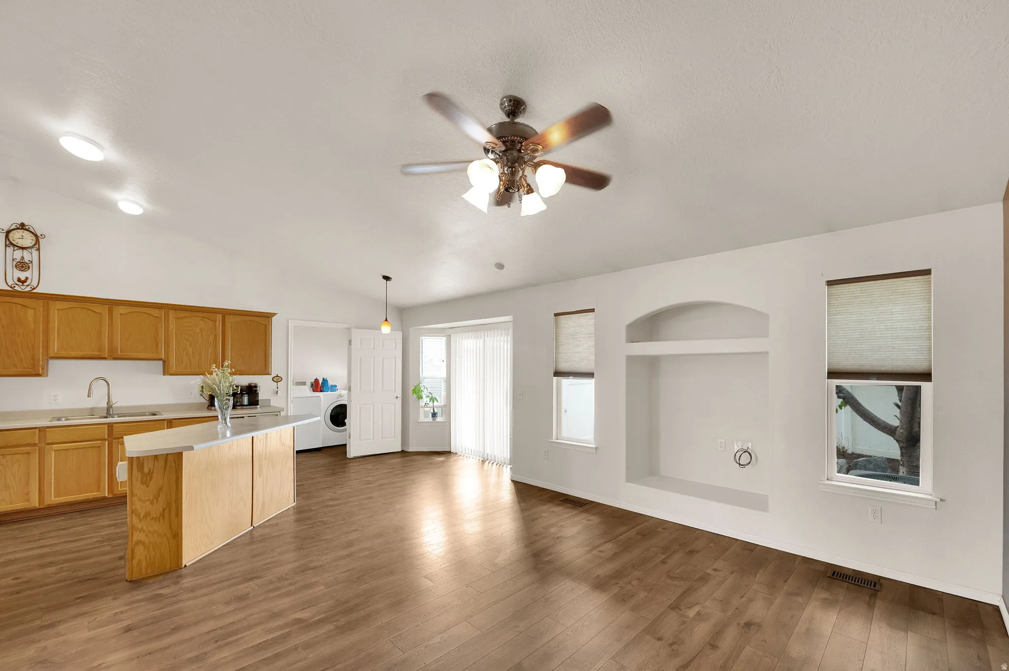 Vaulted ceiling with a ceiling fan light fixture, wood-finish flooring, kitchen island with a built-in sink, sliding glass door, and a built-in media niche