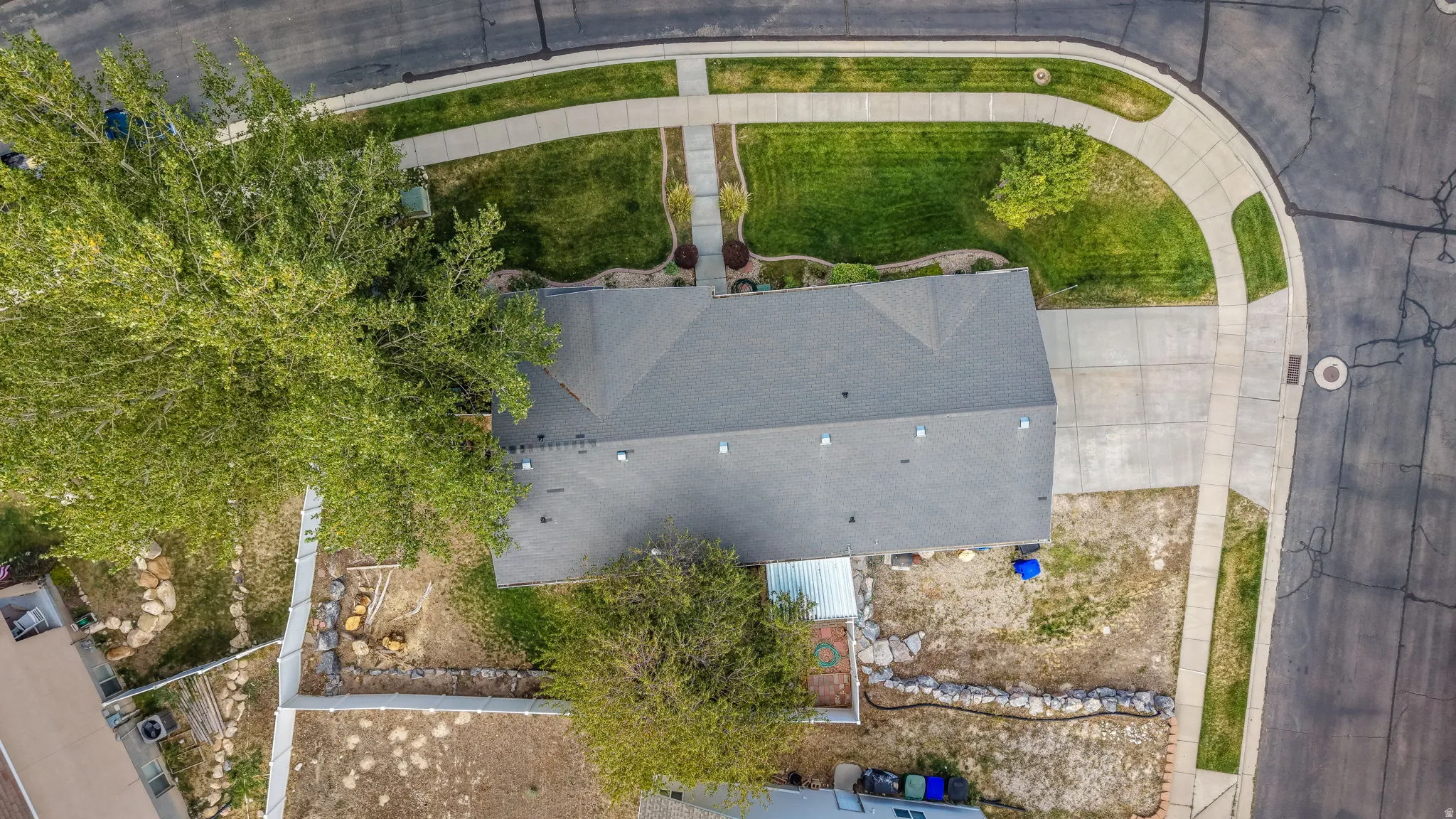 Asphalt shingle roof, expansive paved driveway, concrete sidewalks, and mature trees