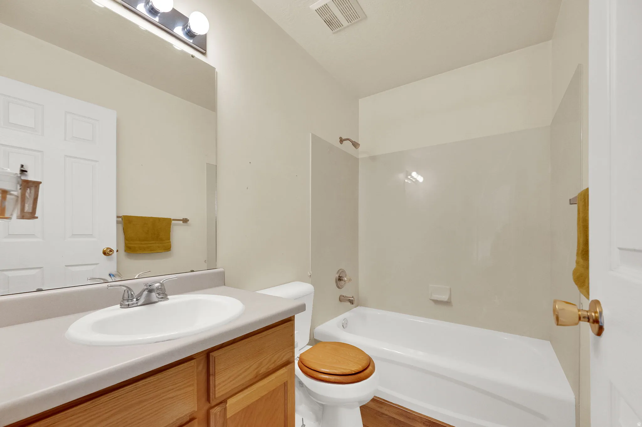 Vanity with integrated sink, light wood-finish cabinetry, full-width wall mirror, and a white bathtub with shower