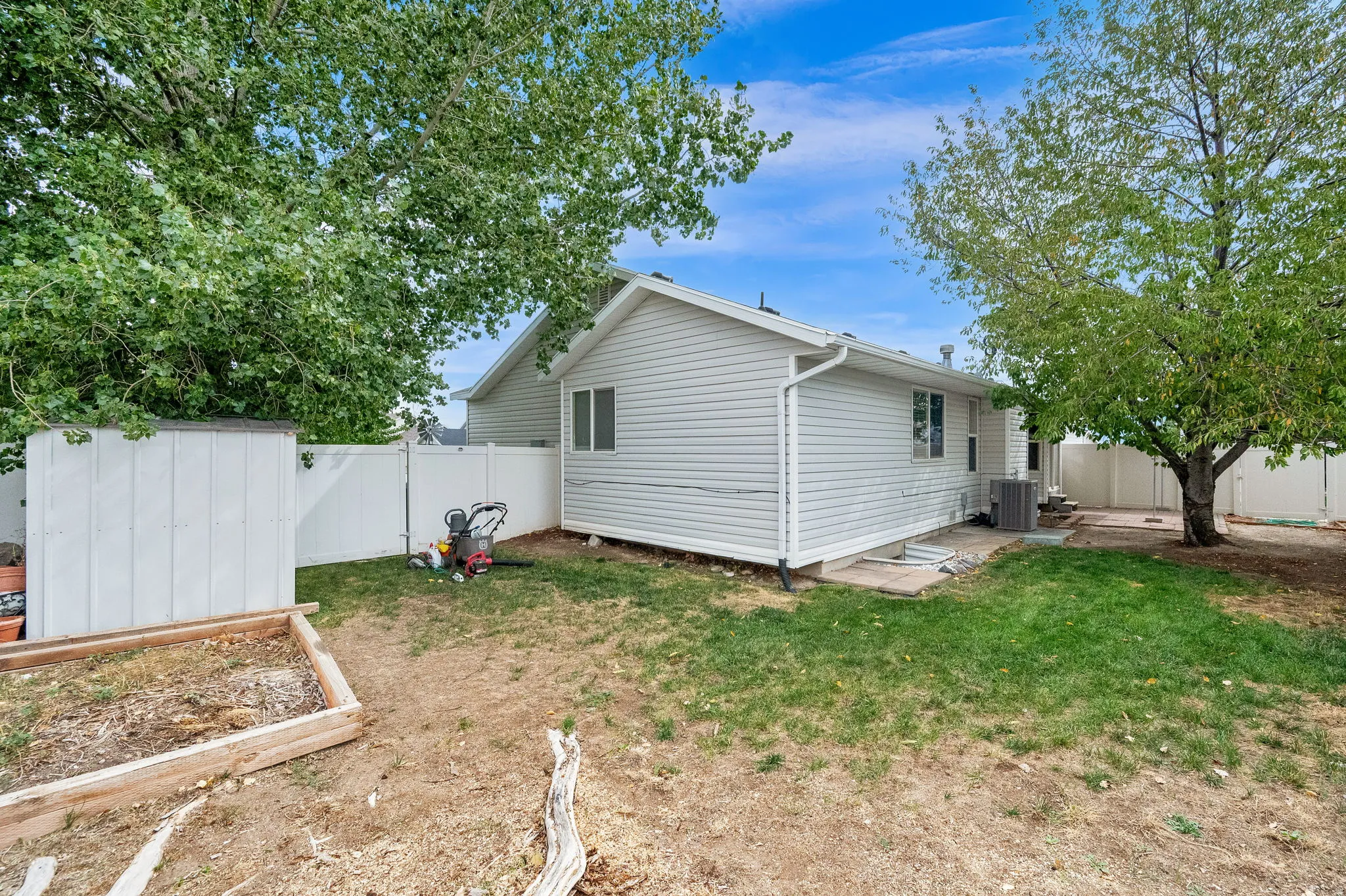 Expansive backyard featuring a white vinyl fence, mature trees, a garden bed, and a storage shed
