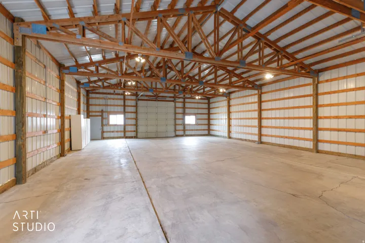 Expansive interior space featuring concrete flooring, exposed wood beam construction, corrugated metal walls, multiple overhead lighting fixtures, and a large roll-up door