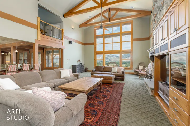 Vaulted ceiling with exposed wood beams, expansive window wall, built-in cabinetry, view of kitchen and upper-level game room handrail.