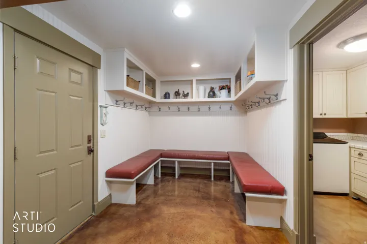 Built-in bench seating with upholstered cushions, wall-mounted coat hooks, overhead shelving, recessed lighting, and stained flooring surface.