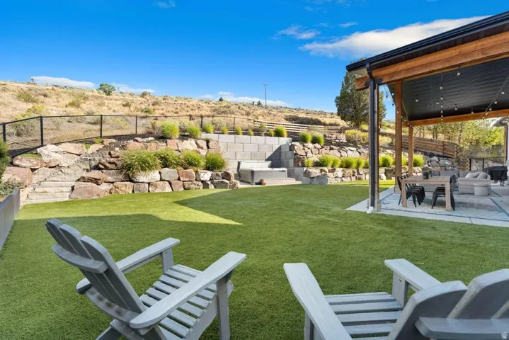 Expansive artificial turf lawn, covered patio with wood beams, built-in spa with stone and block retaining walls, and black metal fencing