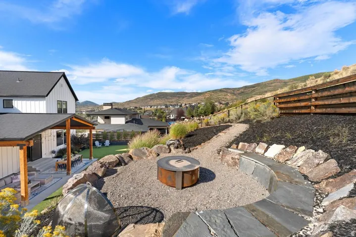 Exterior featuring a modern farmhouse aesthetic, covered patio with wood beams, a gravel pathway, a built-in fire pit, and a natural stone retaining wall