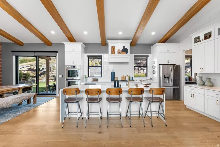Expansive kitchen featuring wood-finish flooring, exposed ceiling beams, a large central island, white cabinetry, and stainless steel appliances