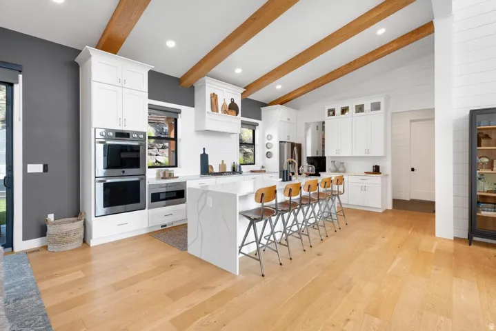 Kitchen featuring vaulted ceilings with exposed wood beams, wood-finish flooring, stainless steel double ovens, and a center island with a waterfall countertop