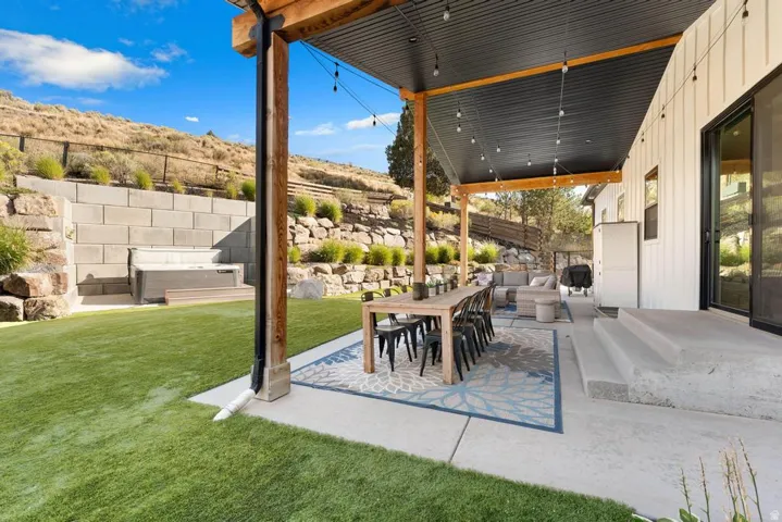 Covered patio with exposed wood beams, concrete patio, artificial turf, and retaining wall