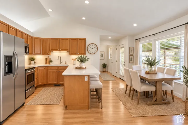 Vaulted ceilings with recessed lighting, wood-finish flooring, a kitchen featuring wood cabinetry, stainless steel appliances, a kitchen island with a light-toned countertop, and an adjacent dining area with large windows