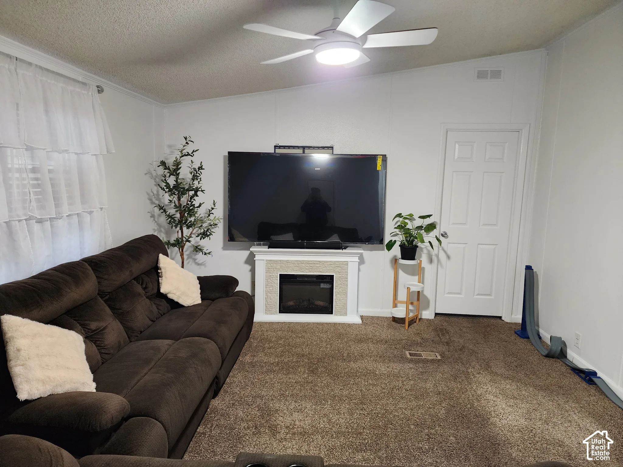 Electric fireplace with a decorative mantel, ceiling fan with integrated lighting, neutral carpeting, and a six-panel interior door