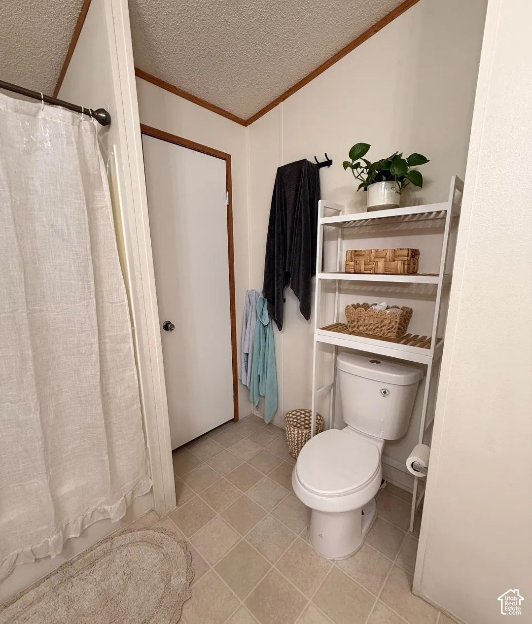 White toilet with an over-the-toilet shelving unit, light-toned square floor tiles, and a privacy door