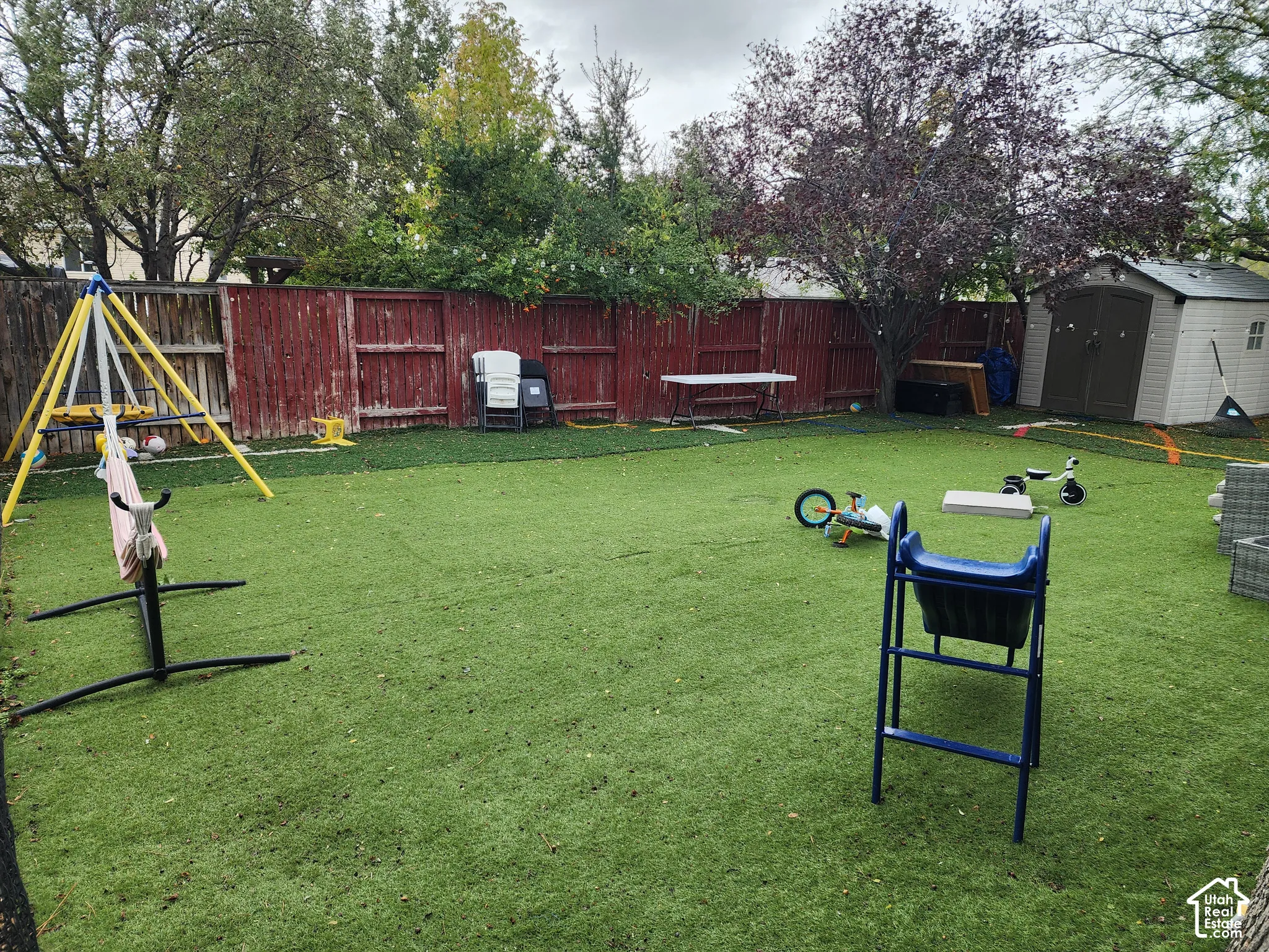 Fenced backyard featuring synthetic turf, mature trees, and a detached storage shed