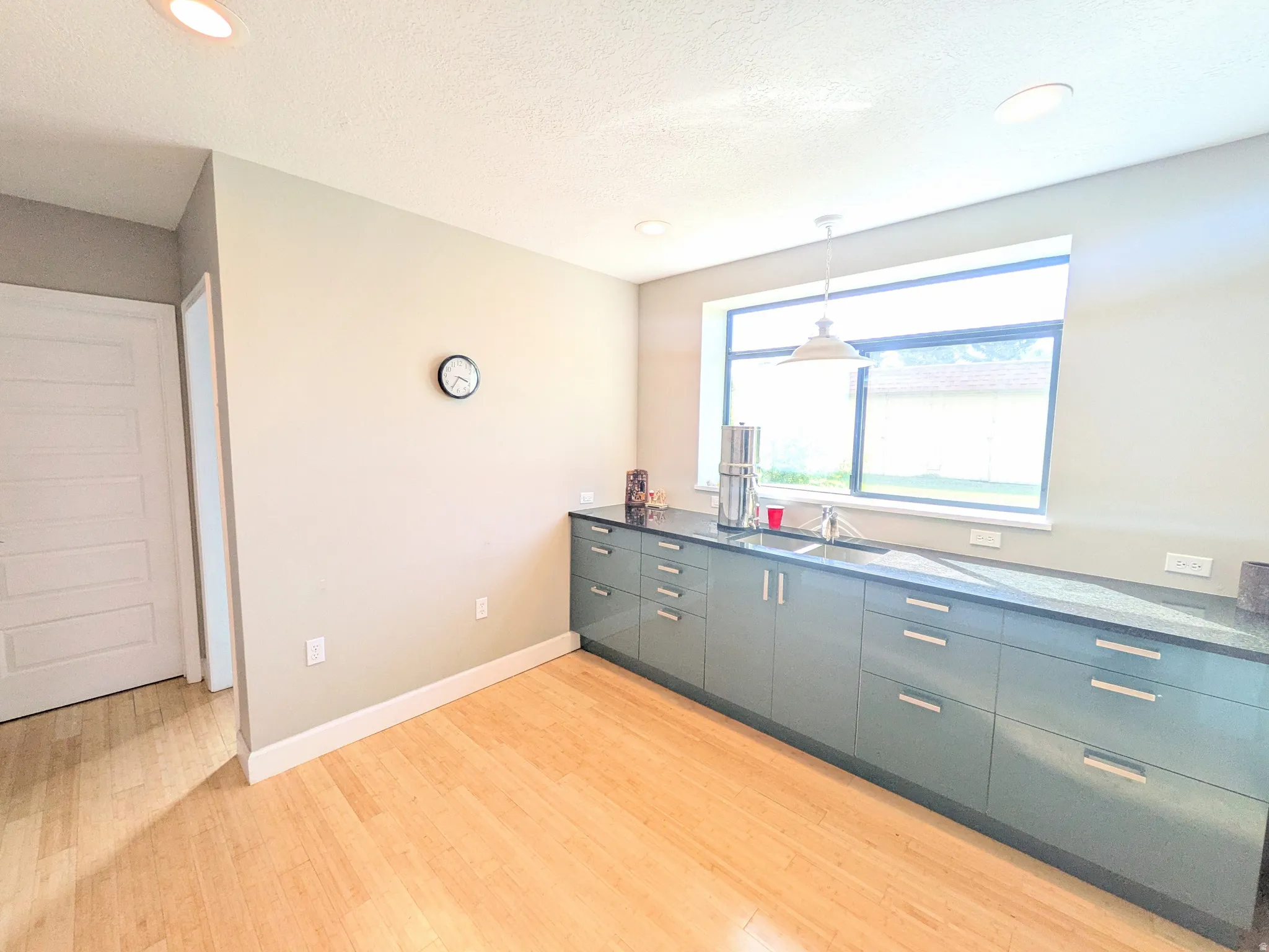 Wood-finish flooring, extensive lower cabinetry with linear hardware, dark countertops, and a large window with pendant lighting