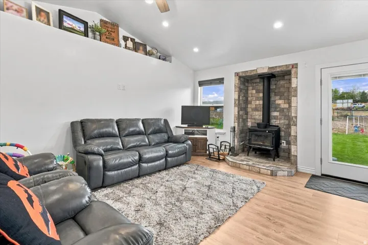Vaulted ceiling with recessed lighting, wood-finish flooring, stone-clad wood-burning stove with hearth, window with roller shade, and glass-paneled exterior door