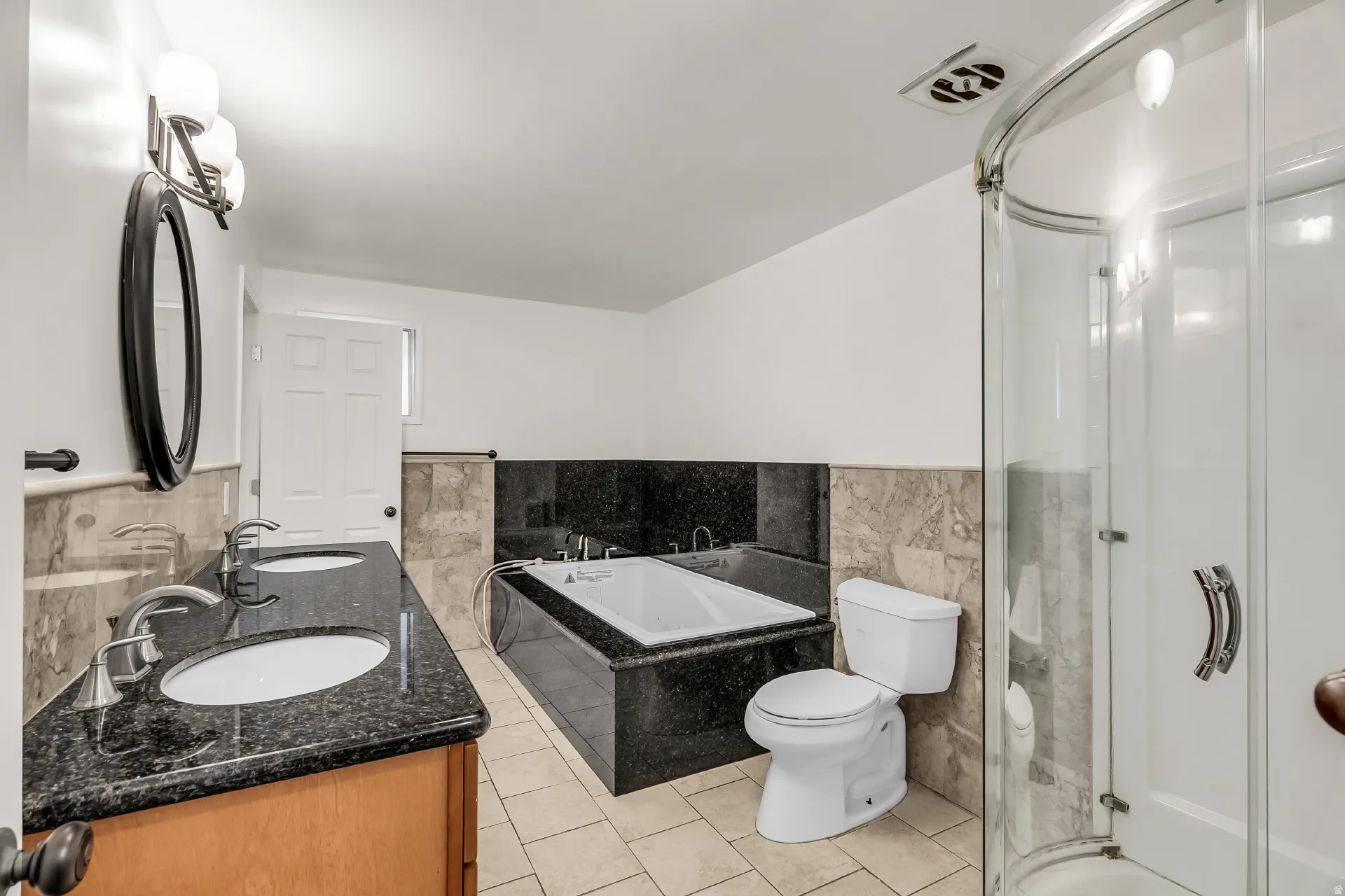 Dual vanity with dark granite countertops, built-in soaking tub with dark surround, glass-enclosed shower, and tile flooring