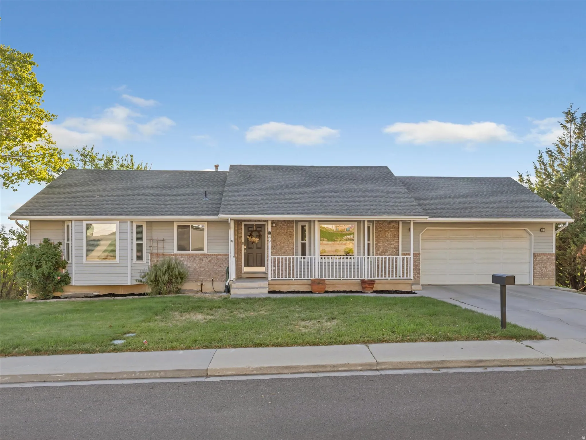 Light gray siding exterior with stone accents, a dark gray shingle roof, an integrated garage, a covered front porch with white railings, and a concrete driveway