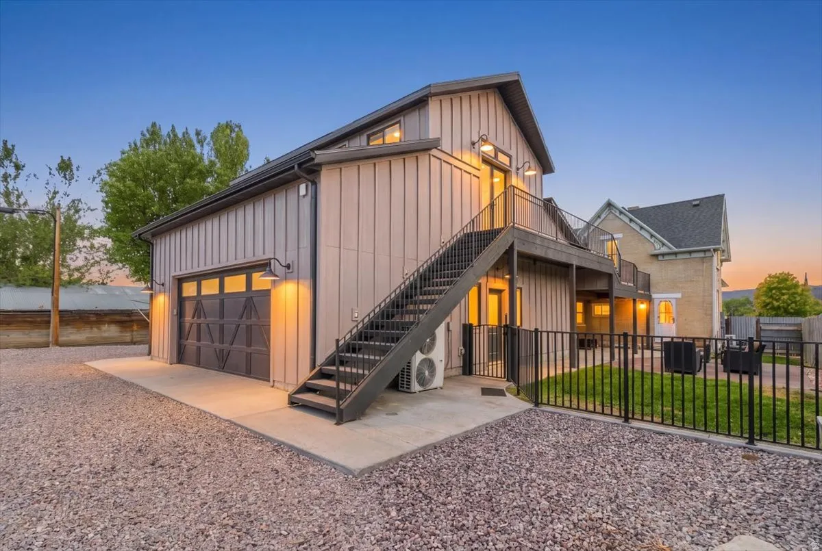 Modern industrial residence featuring corrugated metal siding, expansive garage with overhead doors, external metal staircase with railing, warm interior lighting, and manicured gravel landscaping with mature trees and black perimeter fencing