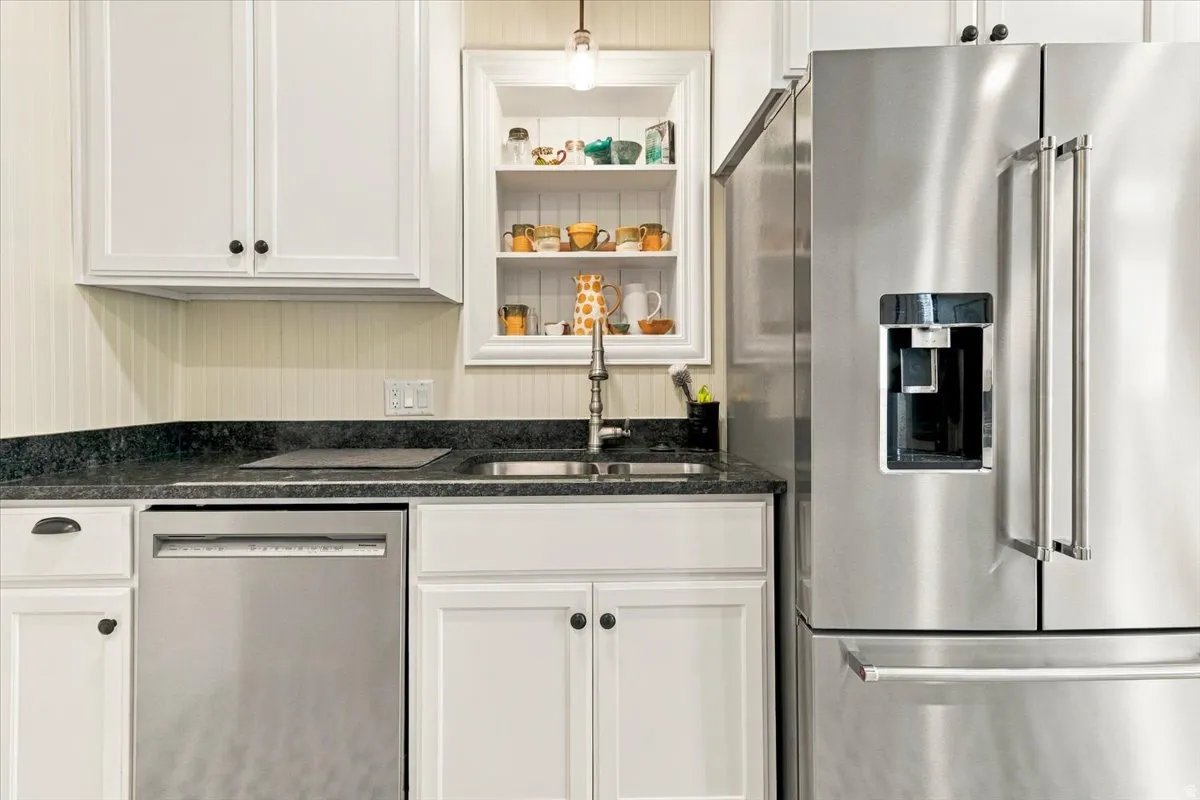 Stainless steel French door refrigerator, white shaker-style cabinetry, black countertop, stainless steel dishwasher, and a built-in recessed shelf