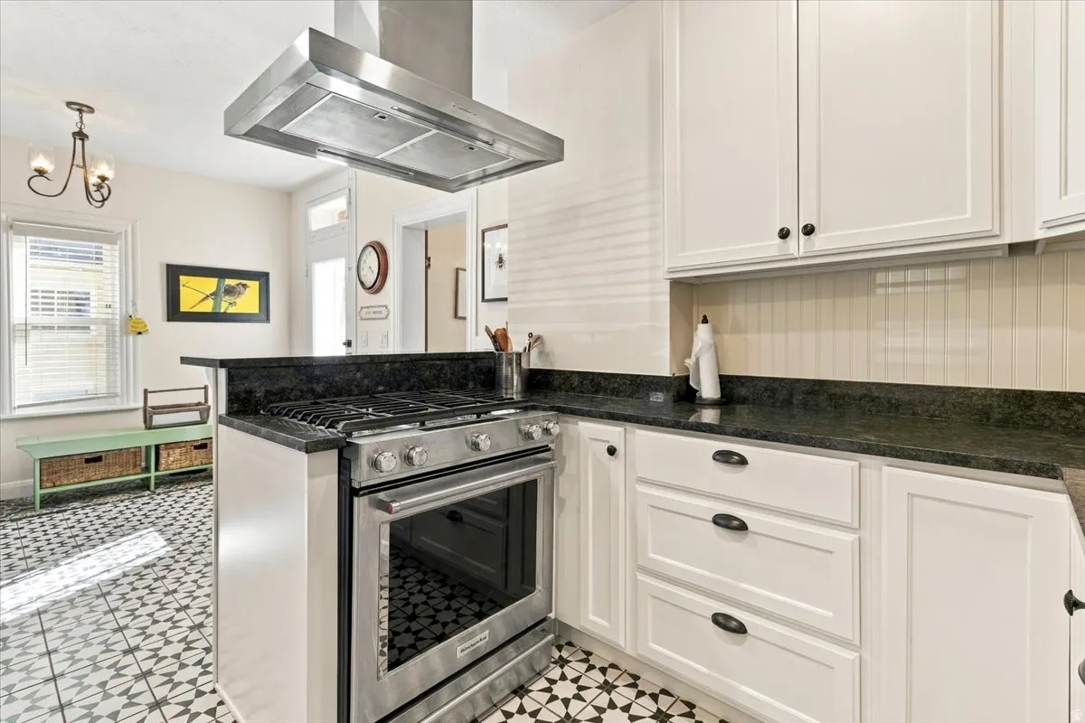 Stainless steel range with coordinating range hood, white shaker-style cabinetry, dark stone countertops, and patterned tile flooring