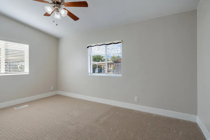 Neutral tone wall paint with white trim, tan carpet flooring, two windows with horizontal blinds, and a ceiling fan with integrated lighting