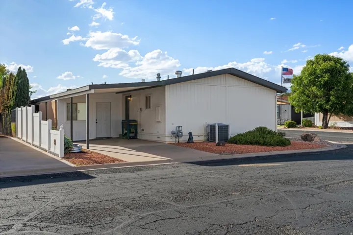 White exterior siding with a carport, paved driveway, privacy fence, mature landscaping, and a compact HVAC unit
