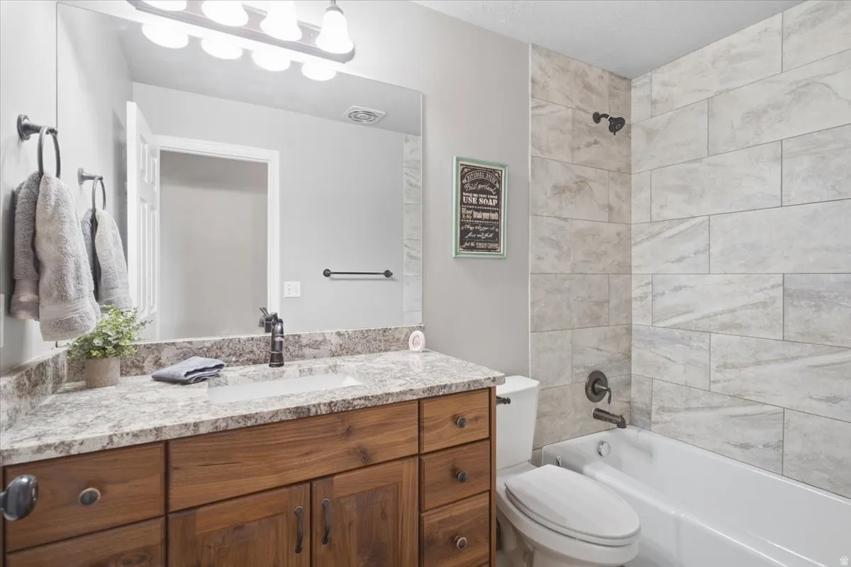 Bathroom featuring a wood-finish vanity with an integrated sink, a speckled countertop, a built-in tub with a shower, and large format wall tile