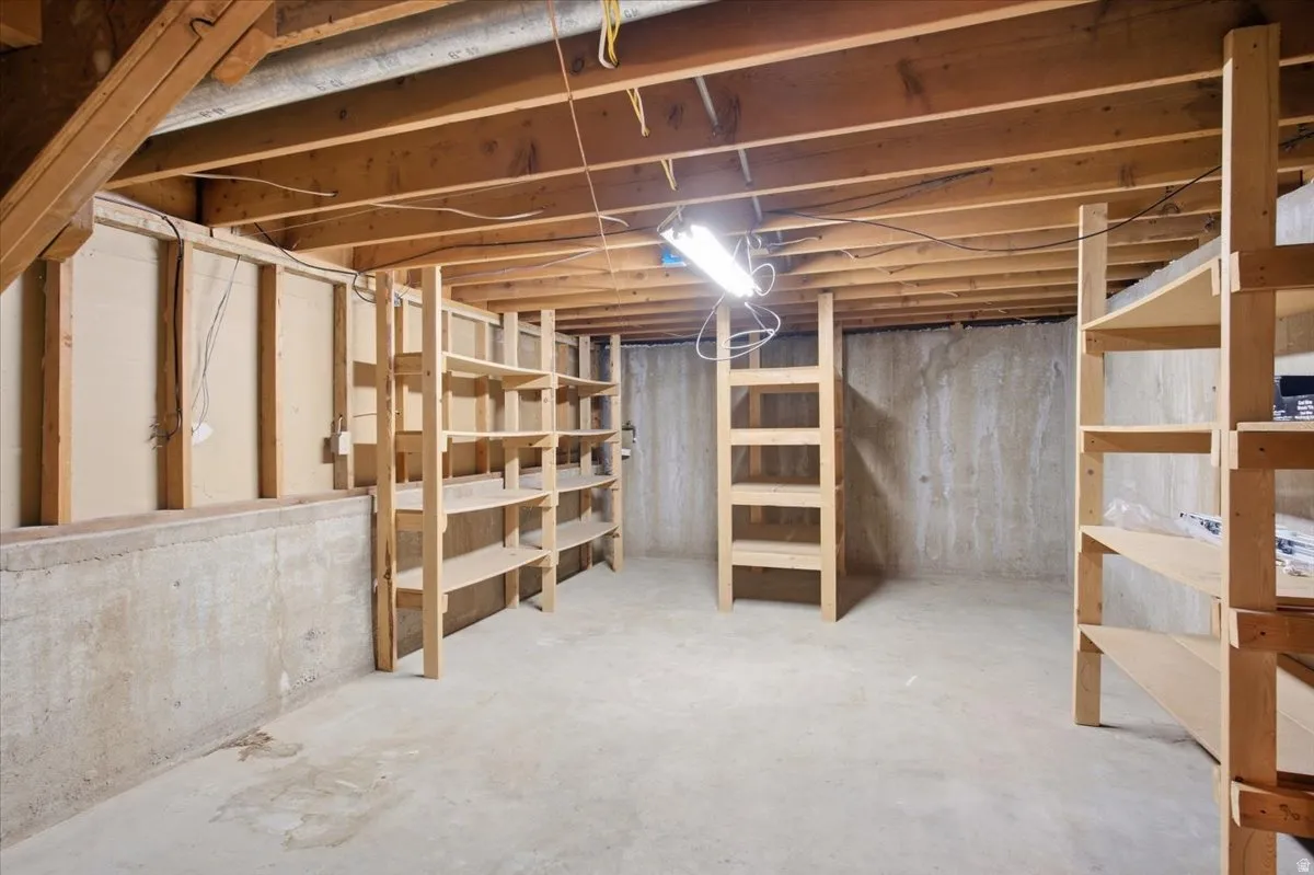 Unfinished basement featuring exposed wood joists, concrete foundation walls, concrete flooring, and built-in wood shelving units