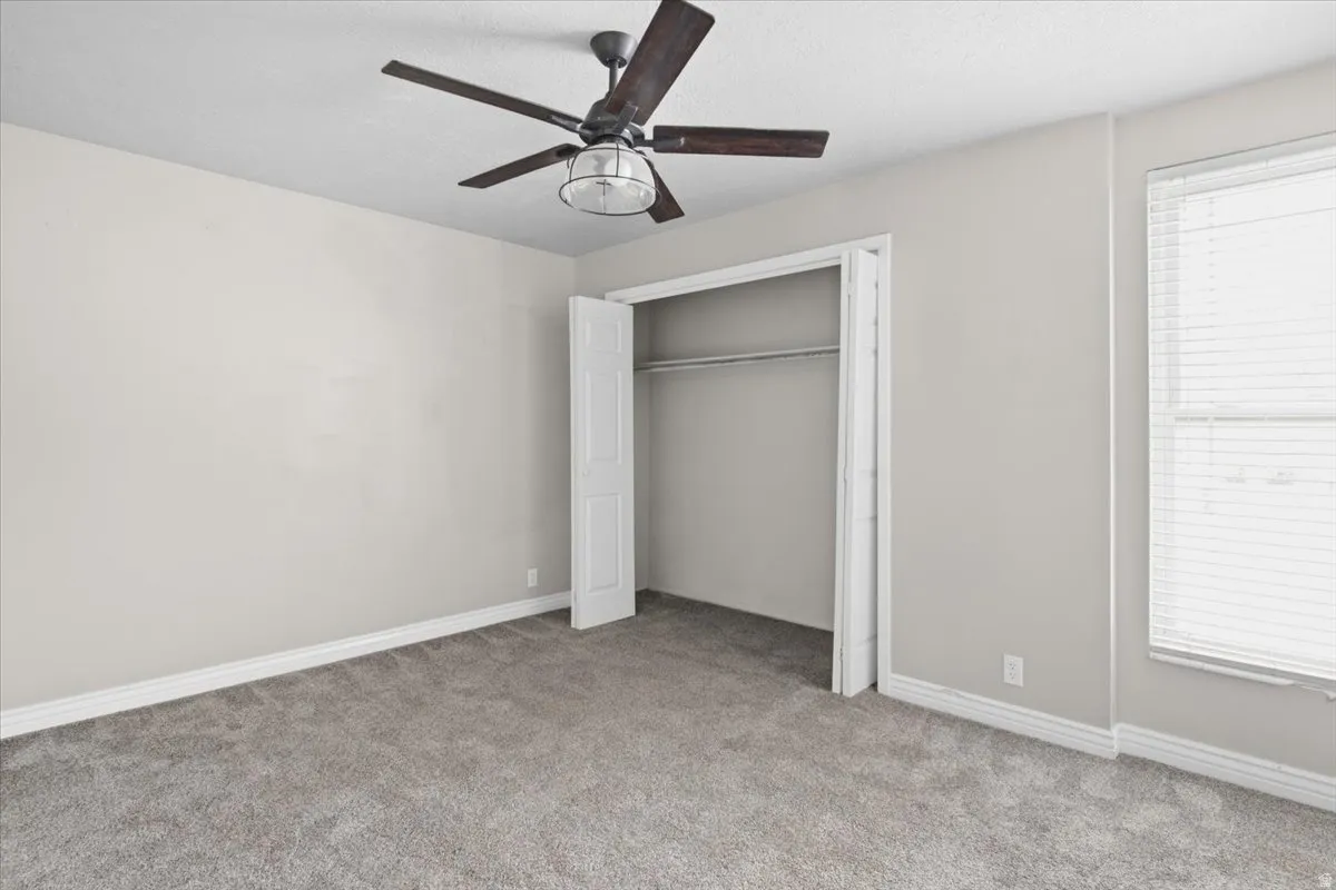 Soft gray carpeting, neutral wall palette, ceiling fan with integrated lighting, window with blinds, and bi-fold closet doors