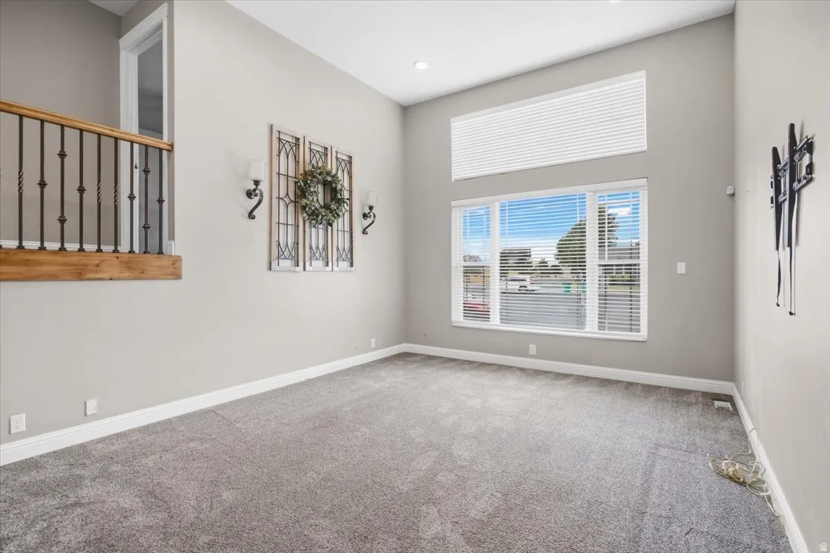 Vaulted ceilings, large windows with blinds, gray carpeting, neutral wall paint, and an upper-level railing with wood and wrought iron details