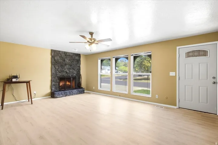 Stone-faced fireplace with hearth, wood-finish flooring, ceiling fan with integrated lighting, multiple windows, and a panel door with an arched glass insert