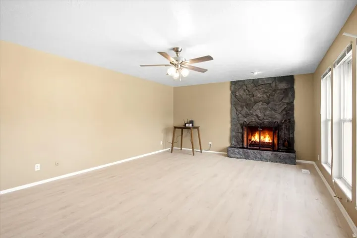 Spacious room featuring wood-finish flooring, a stone-clad fireplace, multiple windows, and a ceiling fan with integrated lighting