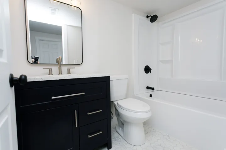 Black vanity with silver hardware, rectangular mirror, white hexagon tile flooring, and integrated shower/tub unit with black fixtures
